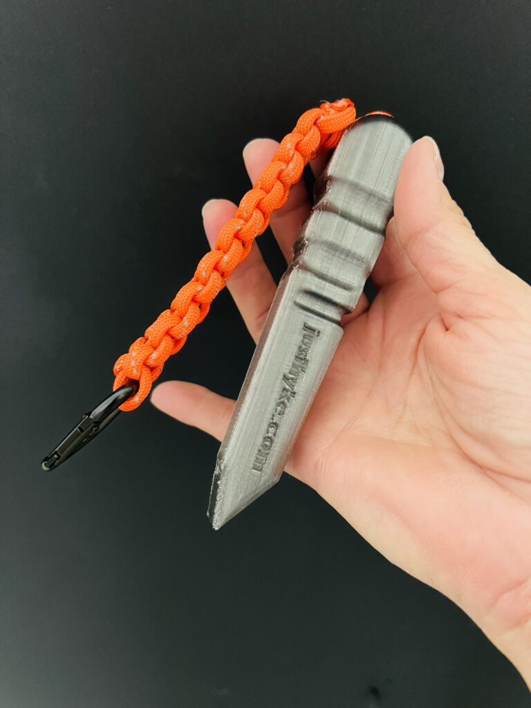 womans small hand holding a grey plastic digging tool with an orange braided paracord and black carabiner.